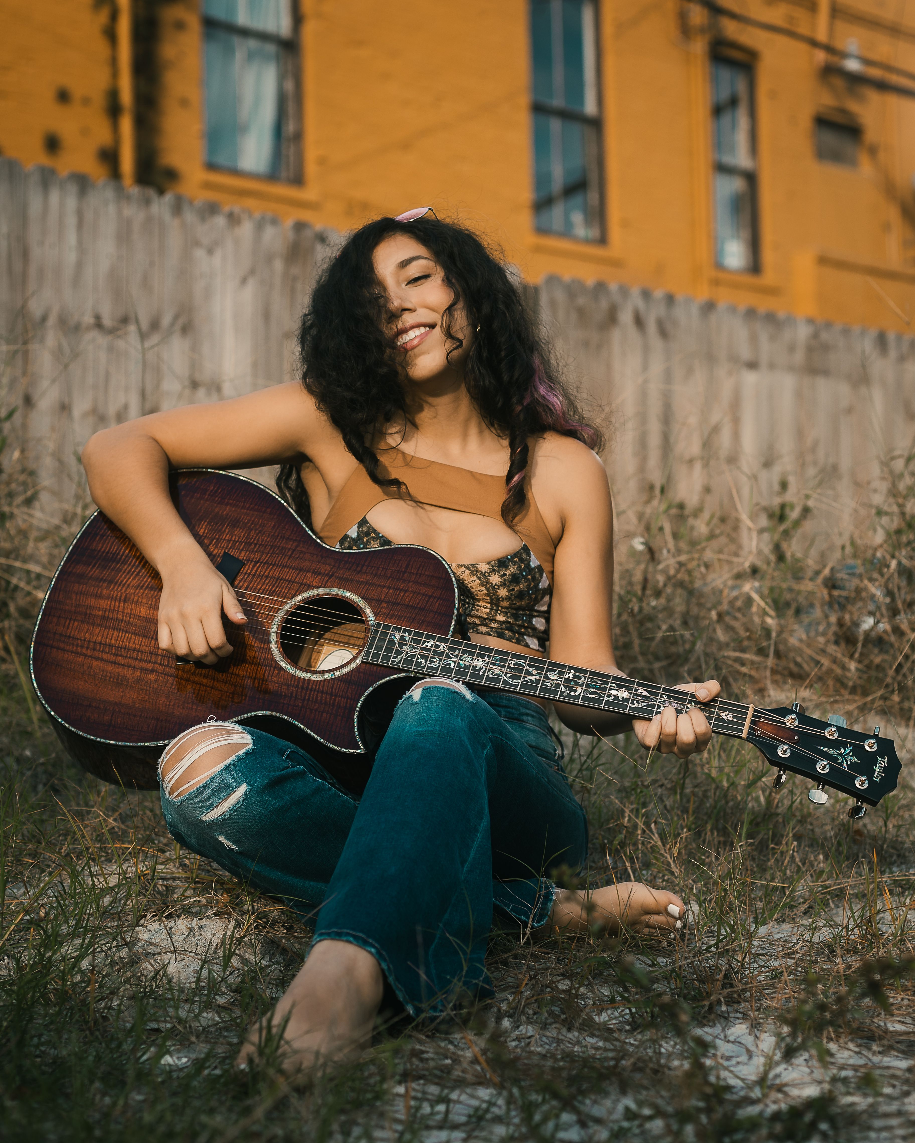A joyful woman with long hair sits barefoot playing guitar against a rustic fence.