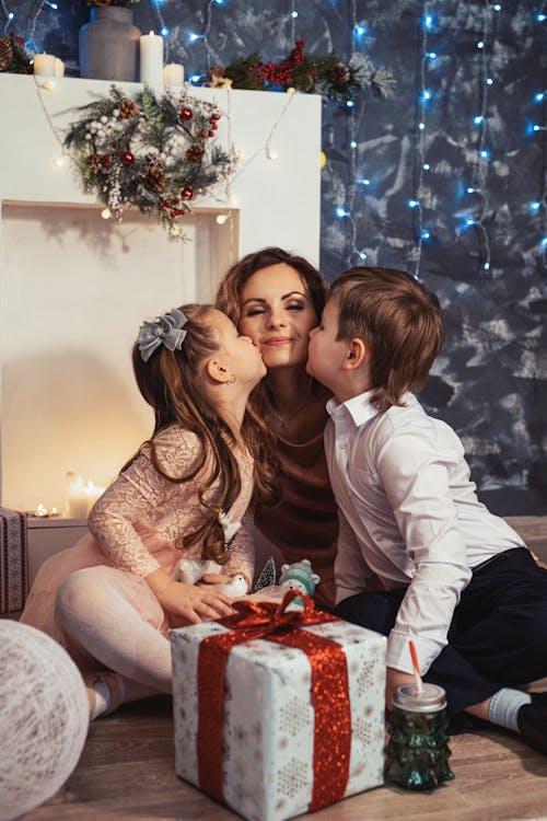 Free Children Kissing their Mother on the Cheeks while Sitting on the Floor in front of a Gift Box Stock Photo