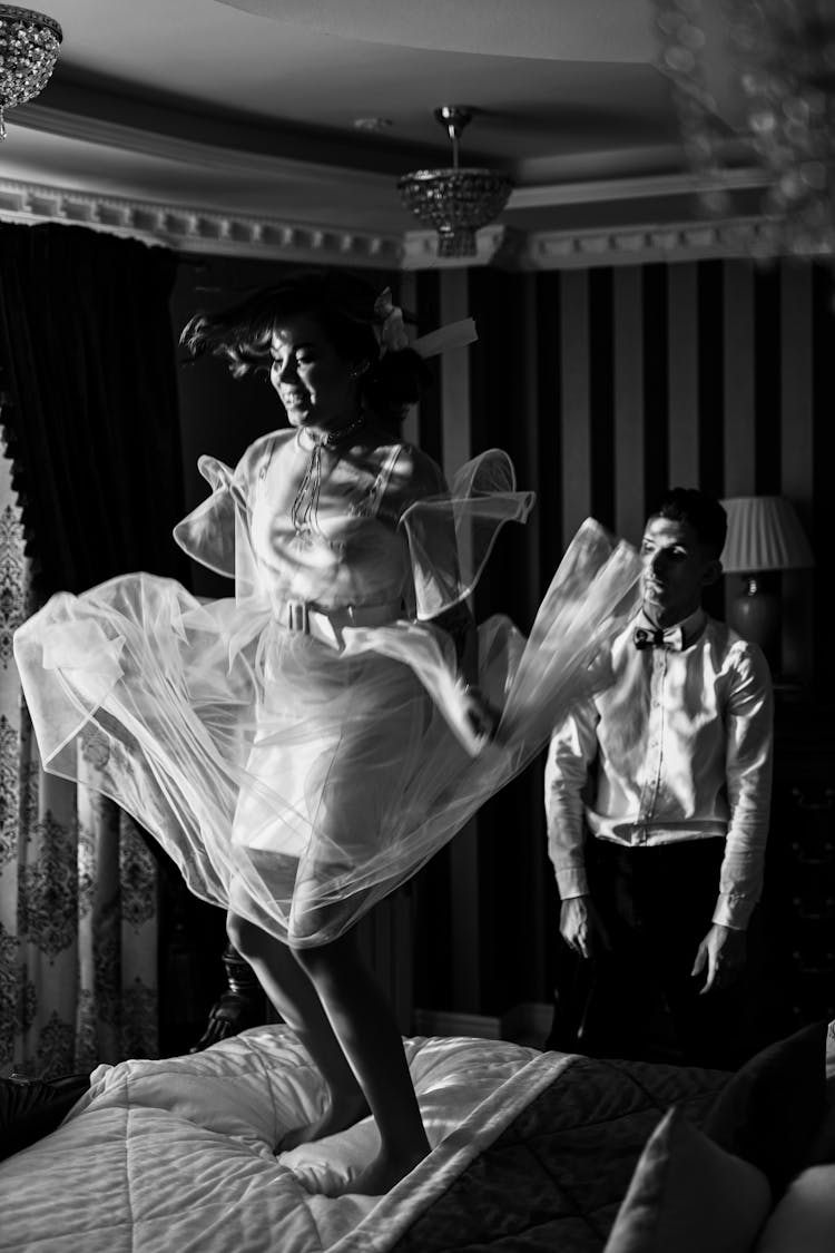 Grayscale Photo Of A Woman Jumping On The Bed Near A Man