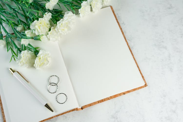 Silver Rings On Top Of A Blank Notebook Beside Pen And White Flowers