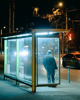 A person stands alone at a bus stop at night, illuminated by city lights and car headlights.