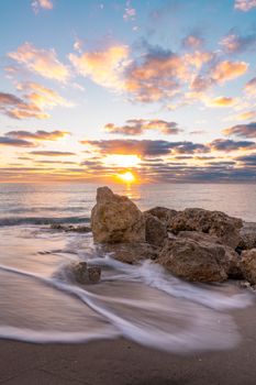 Peaceful sunset over the ocean with rocks and gentle waves in the foreground.