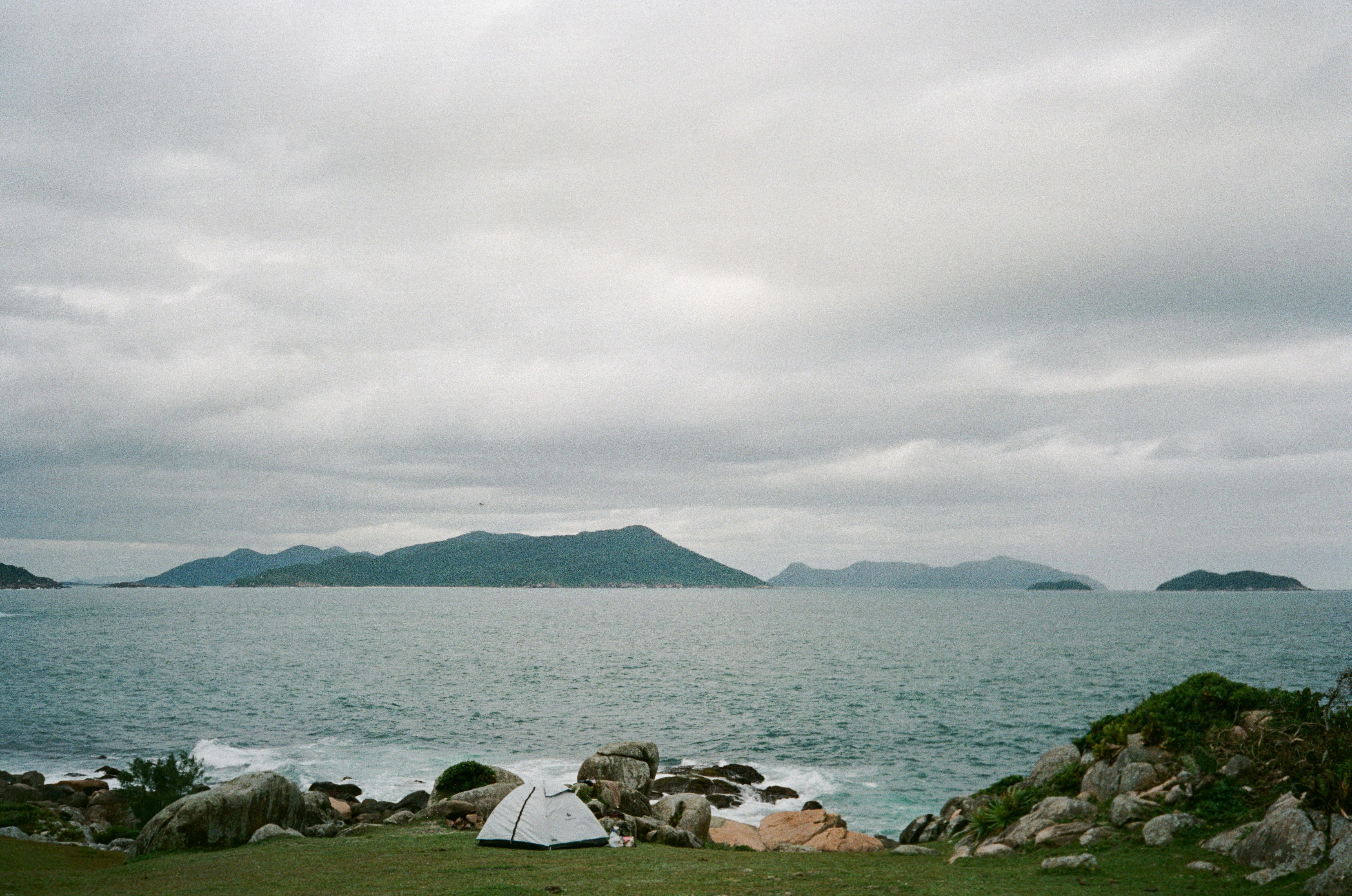 A tranquil camping scene by the sea with rocky landscape and distant islands under a cloudy sky in Santa Catarina, Brazil.