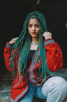 Portrait of a fashionable woman with green braids, wearing a red jacket, posing indoors.