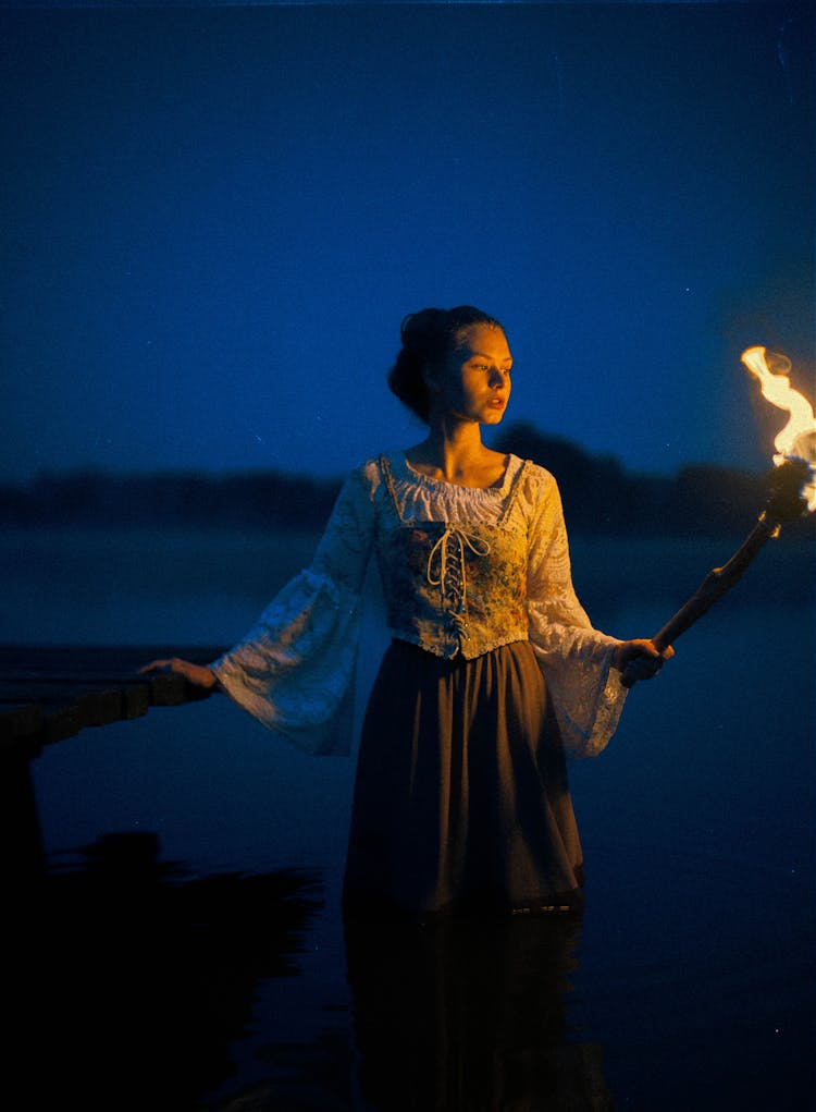 A Woman In Medieval Dress Holding A Torch In The Lake