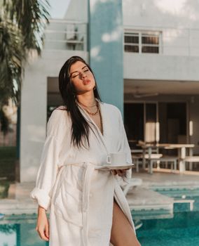 Woman in bathrobe with coffee by pool, enjoying morning sun.