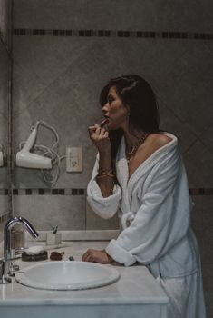 Elegant woman in bathrobe applying lipstick in a modern bathroom.