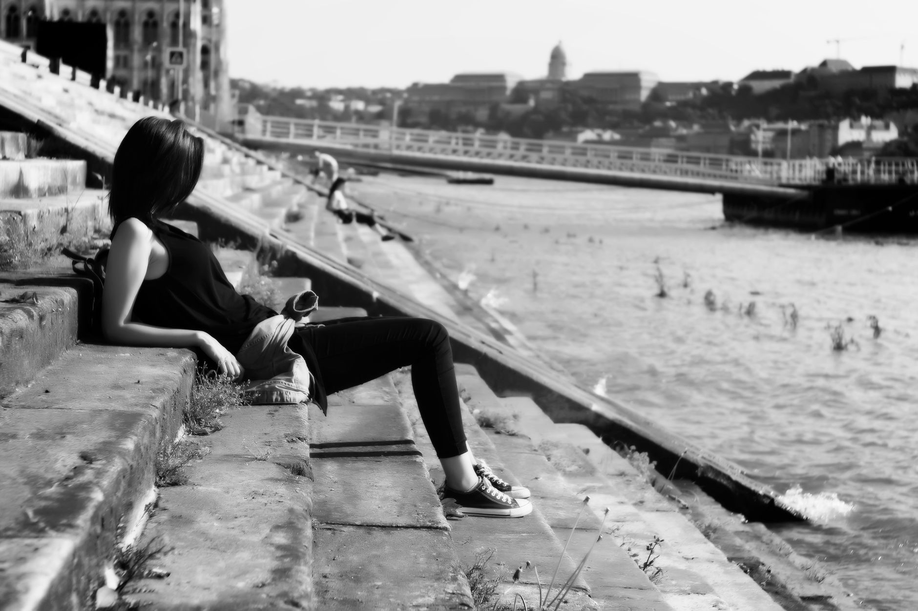 Black and white photo of a woman lounging by a river in an urban area, capturing a serene moment.