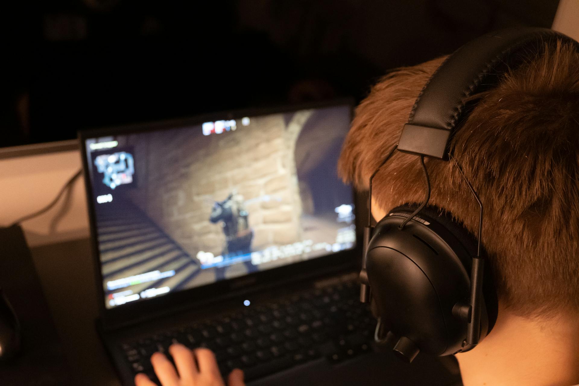 Teenage boy wearing headphones focused on playing a video game on a laptop screen indoors.