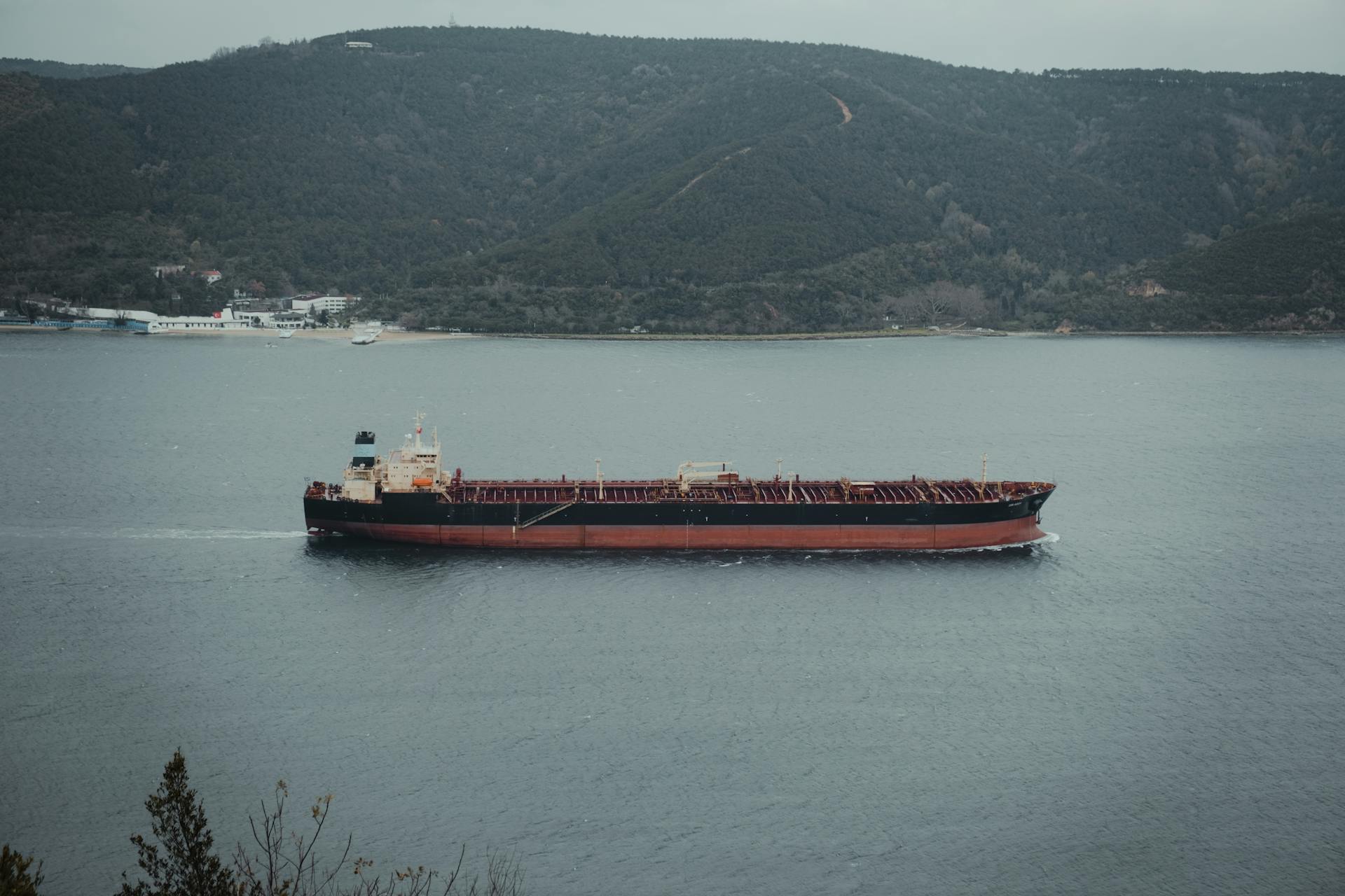 A cargo ship travels smoothly on a wide body of water against a backdrop of lush green mountains.