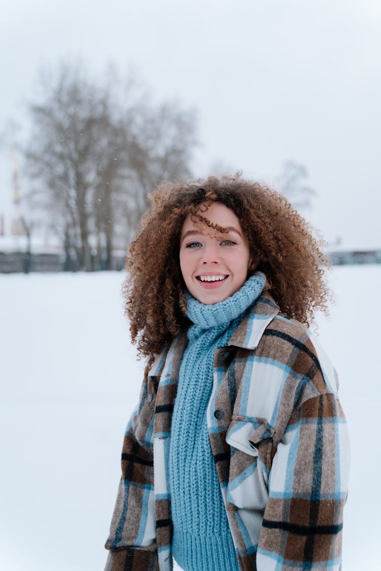 Woman With Curly Hair Wearing Winter Clothes Standing In Snow Field Smiling