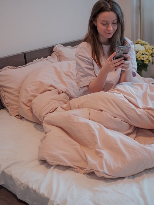 Free A Woman Sitting in Bed Under a Comforter Holding a Cellphone Stock Photo