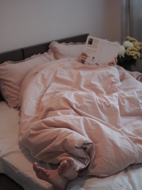 A Person Lying in Bed Under a Pink Comforter Holding a Book