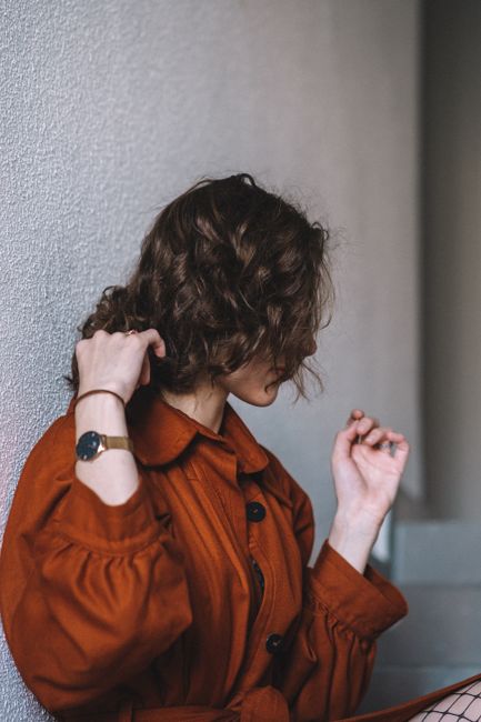 Elegant woman with curly hair in a red coat adjusting her watch indoors against a wall.