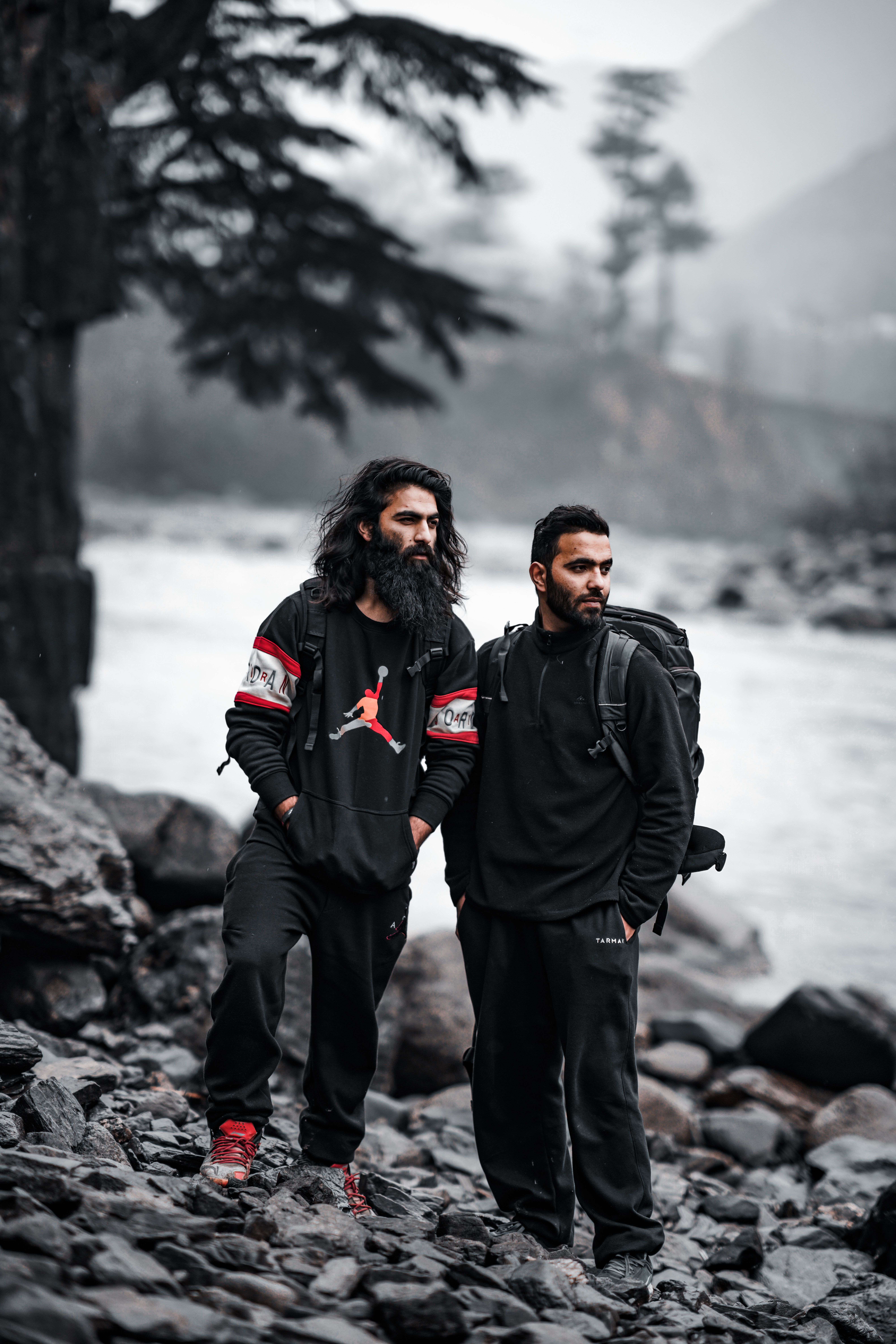 Two men in black clothing exploring a rocky riverbank, surrounded by misty trees.