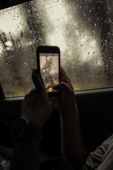 A person uses a smartphone to film raindrops on a car window, capturing the beauty of a rainy day.