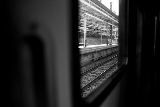 View of a railway station platform and tracks through a train window in black and white.