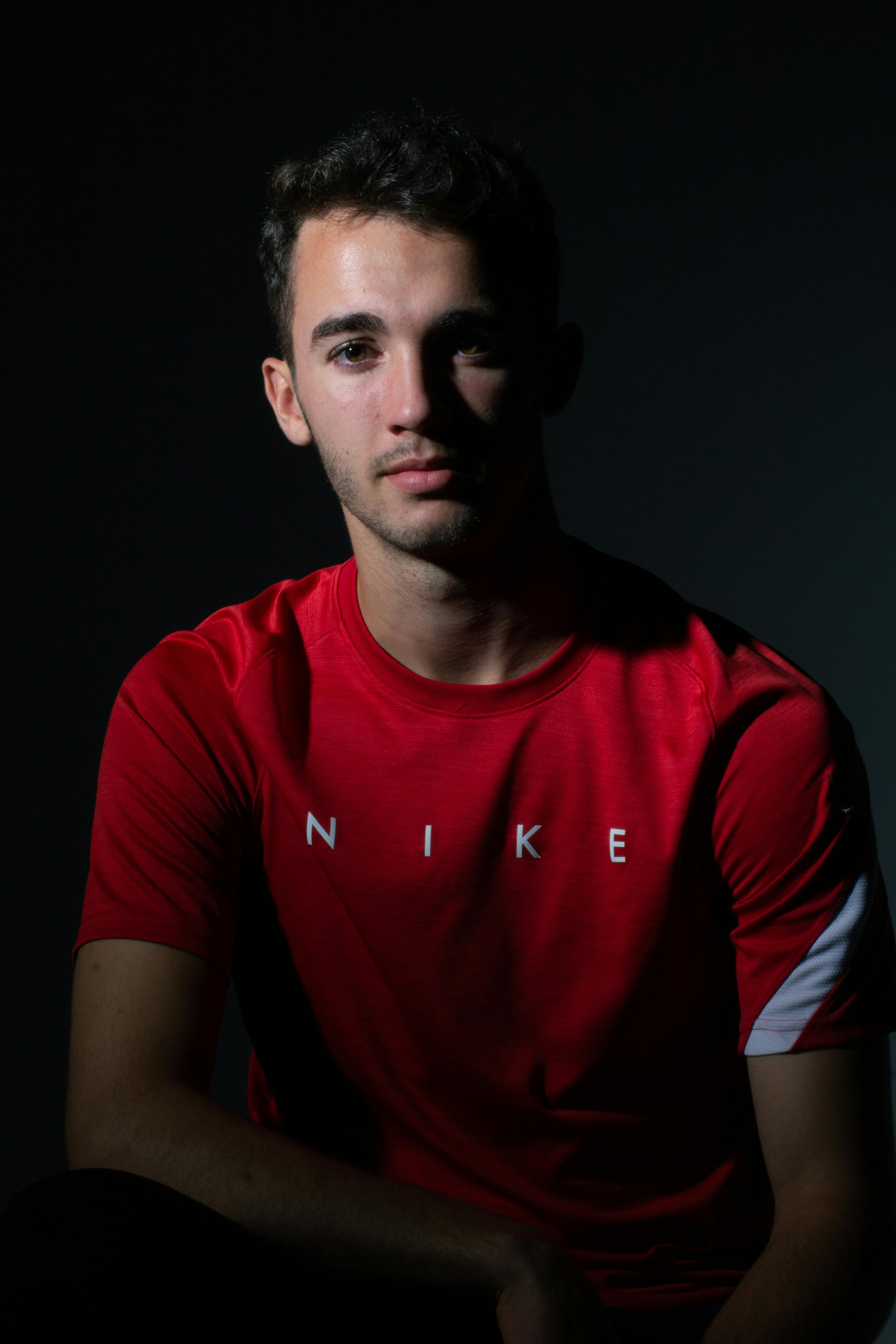 Dramatic studio portrait of a man in a red shirt with shadows and a neutral expression.