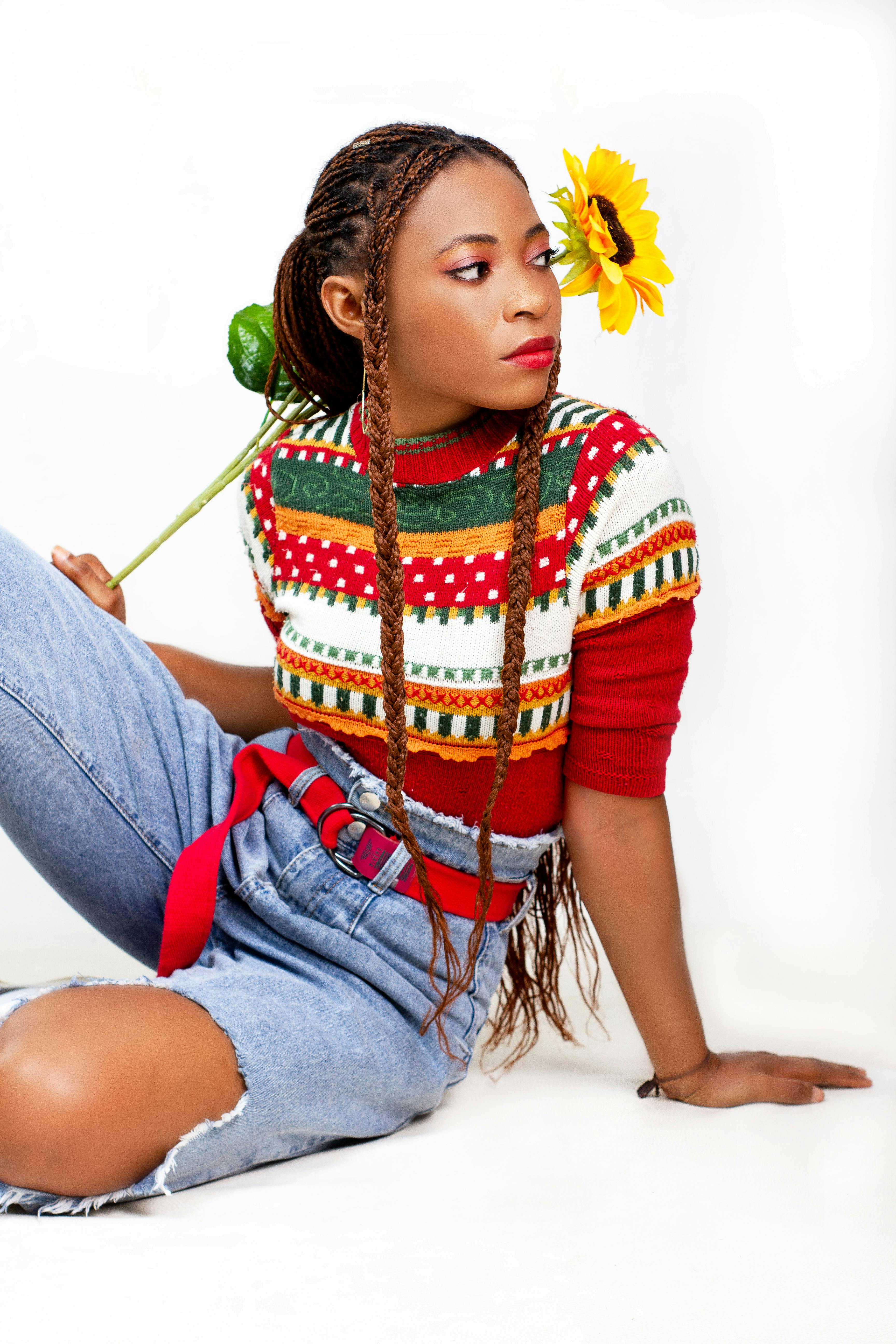 Free Young woman in vibrant sweater and ripped jeans posing with sunflower on white background. Stock Photo