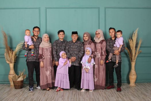 Large family portrait in matching traditional attire, standing together against a teal background. - Fotografer Sidoarjo