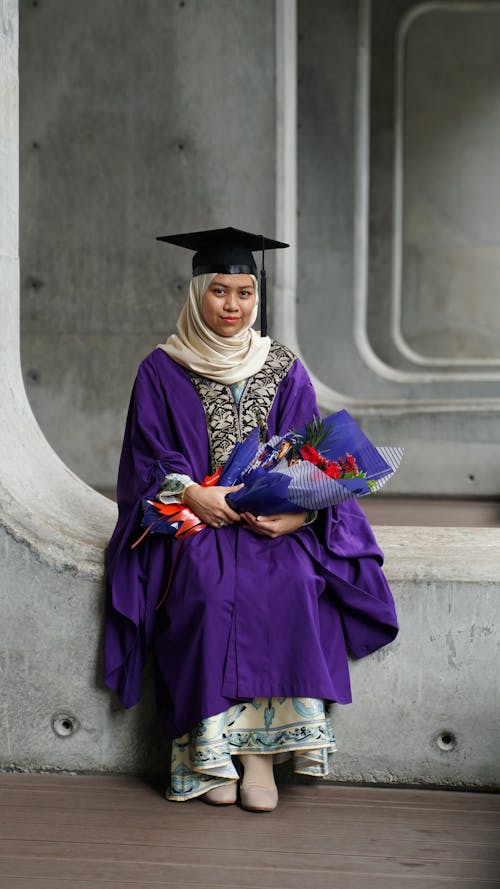 Woman in Purple Academic Gown and White Hijab Sitting on Concrete With Bouquet of Flowers 