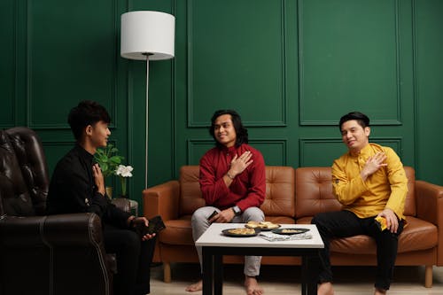 Three Men in Bright Shirts Sitting Barefoot on Brown Leather Couch and Armchair Beside Green Wall