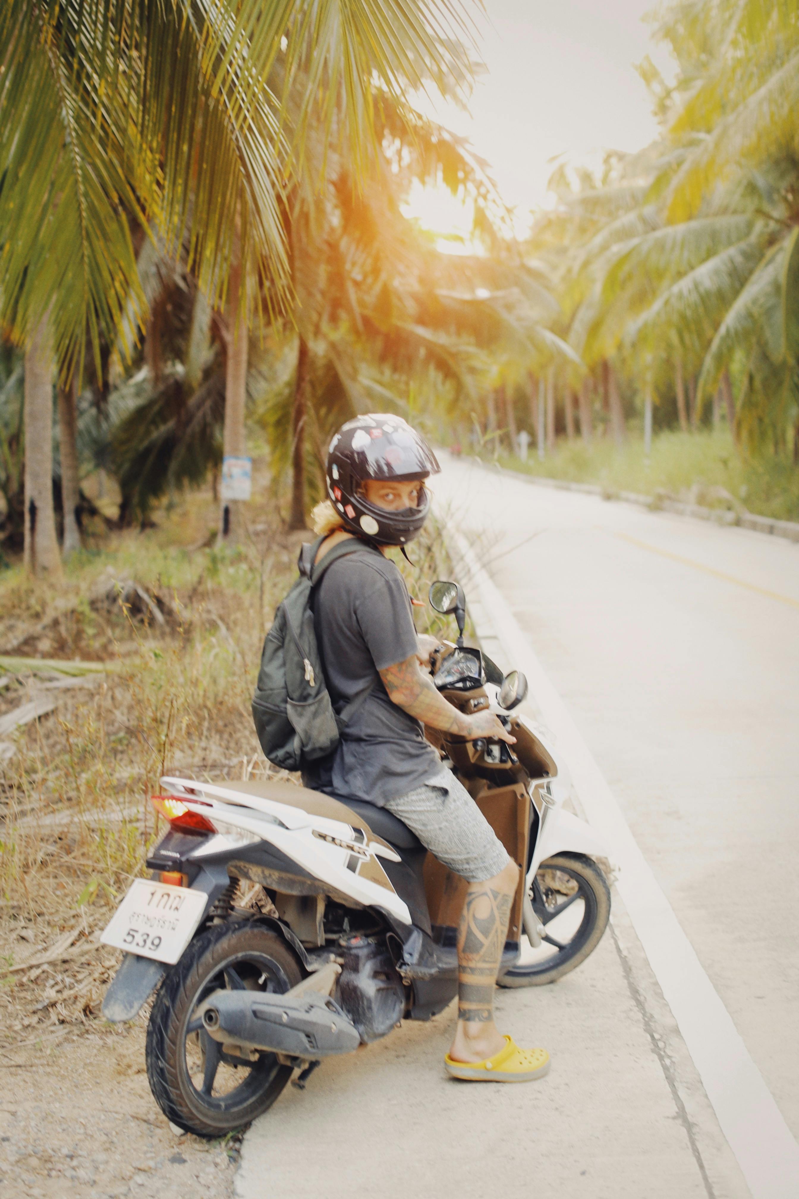 A motorcyclist resting by a tropical road, surrounded by palm trees, in a sunny setting.
