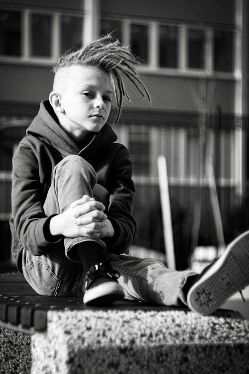 Grayscale Photo of Boy in Hoodie Sitting on Concrete Bench