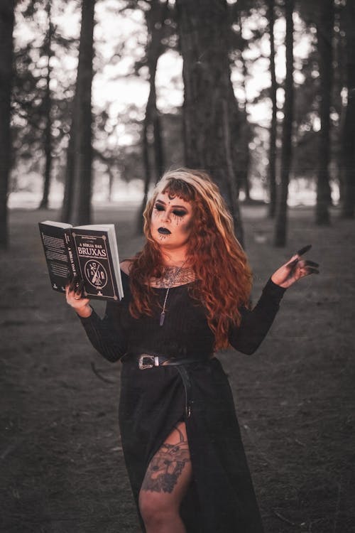 Woman in Black Long Sleeve Dress Standing on Grass Field while Holding a Book