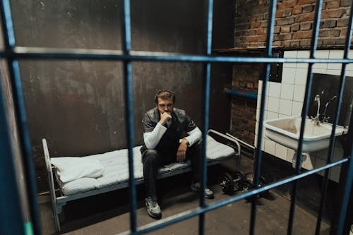 Free A solitary prisoner in a dimly lit cell, sitting on a bed in deep thought, evoking a sense of isolation and introspection. Stock Photo