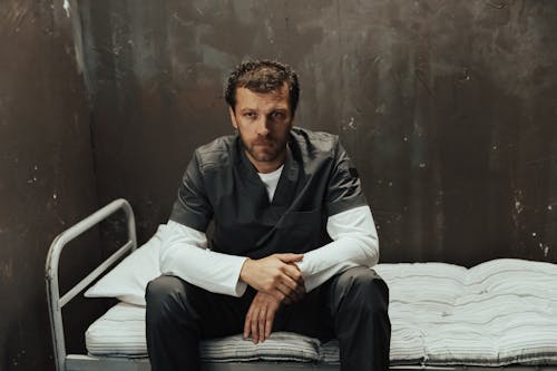 Man in Gray Robe Sitting on Bed in Cell