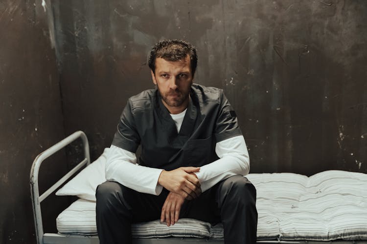 Man In Gray Robe Sitting On Bed In Cell