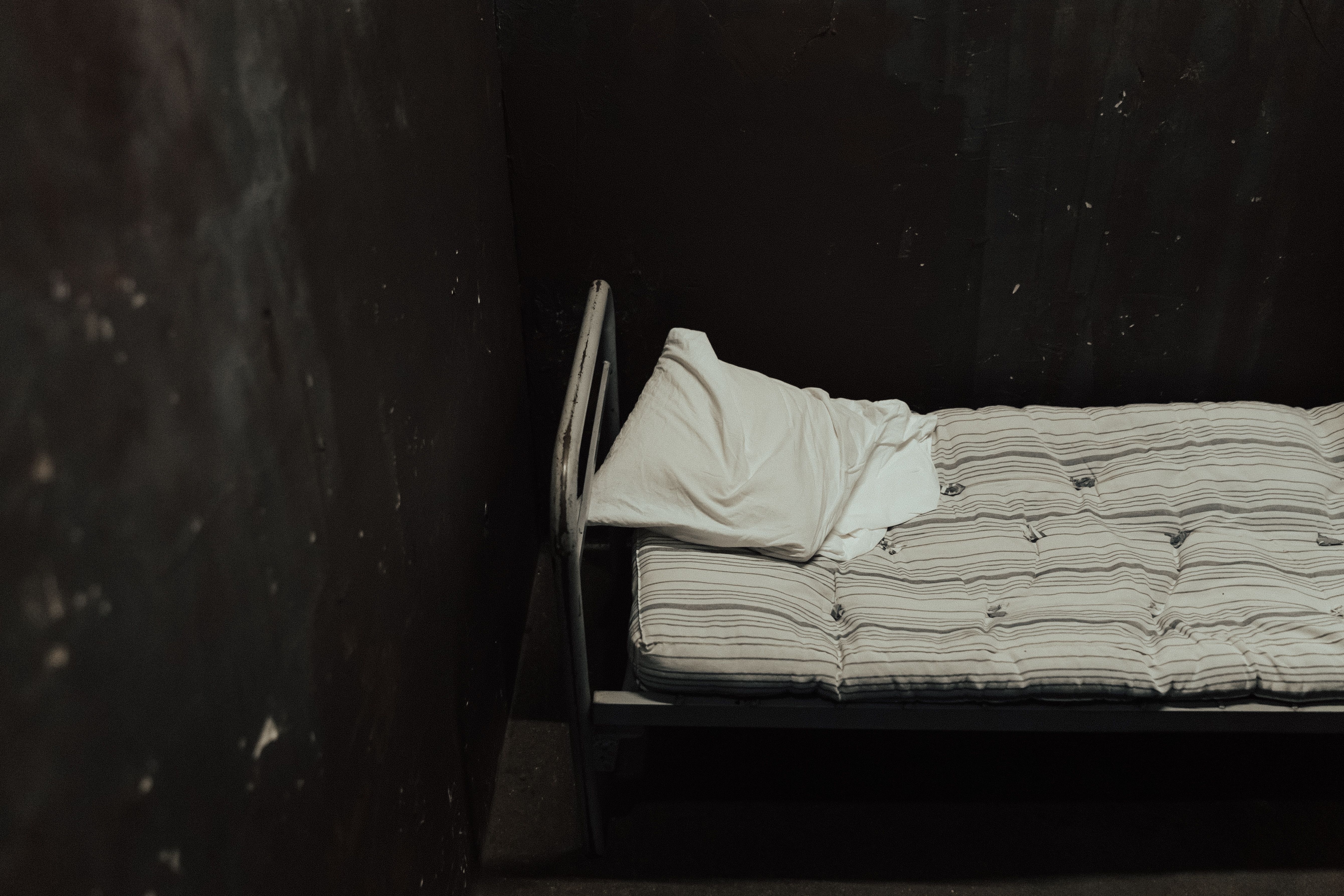 Single bed with striped mattress against a dimly lit wall, creating a somber atmosphere.
