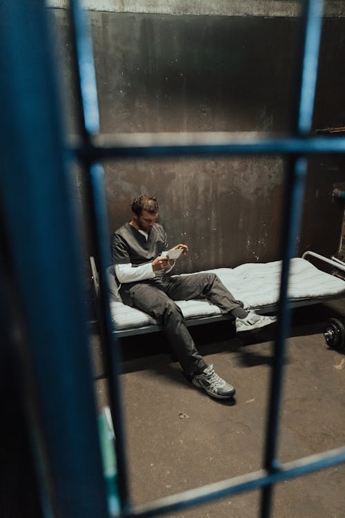 Man in Gray Robe Sitting on Bed and Reading Behind the Bars