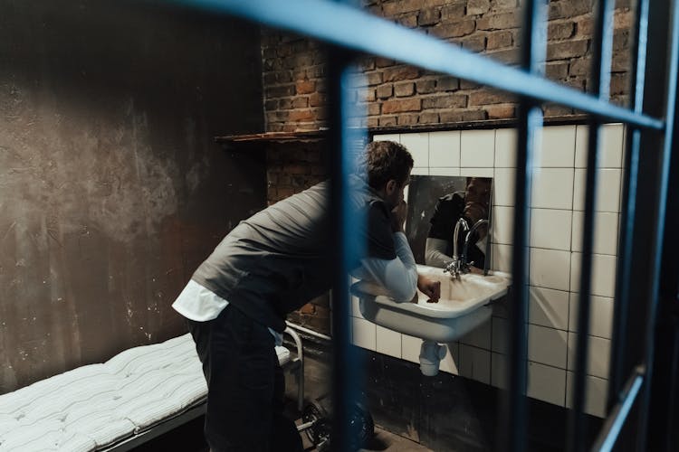 Man In Gray Robe Looking At The Mirror In Cell Behind Bars