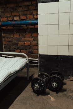 Dumbbells next to a bed in a rustic gym setting with exposed brick wall.
