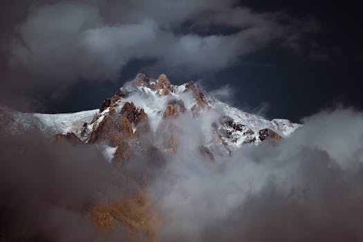 Majestic mountain peak enveloped in clouds with a snowcapped summit under a dramatic sky.
