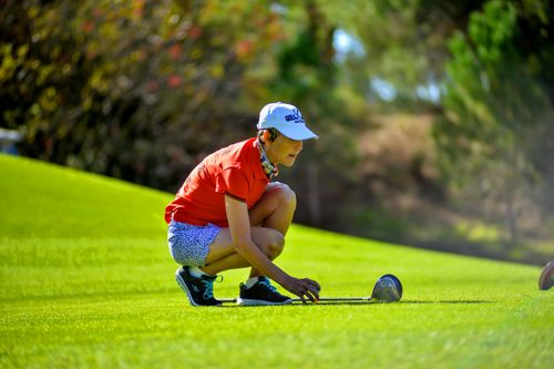 Free Female golfer in red prepares to putt on a lush green course under clear skies. Stock Photo