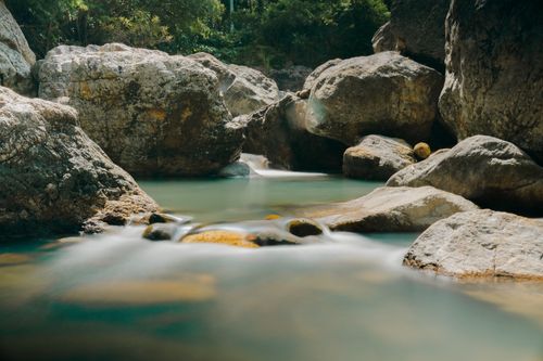 Free A serene river with long exposure flowing through large rocks in a lush landscape. Stock Photo