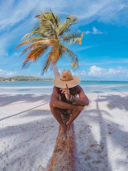 A man with a straw hat sitting on a beach log under a palm tree in Brazil, enjoying a sunny day.