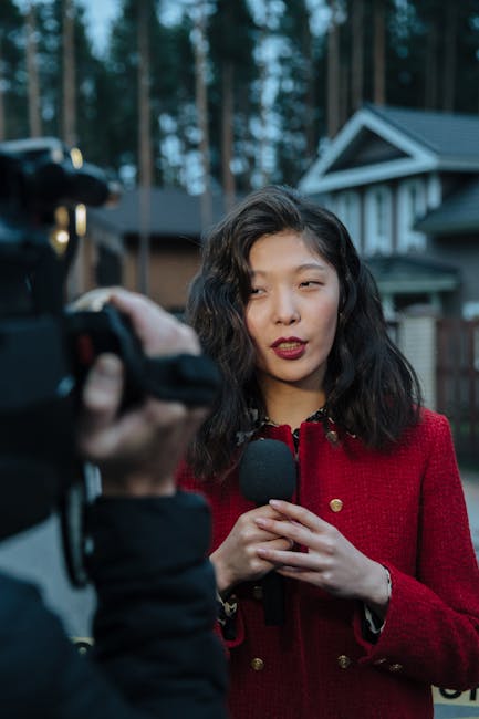 Asian woman reporting news on camera outdoors, wearing red coat.