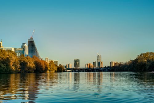 Free City skyline reflecting in the river with a bridge and autumn trees under a blue sky. Stock Photo