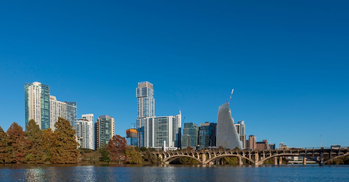 A vibrant city skyline with modern architecture reflecting in the river under clear blue skies.