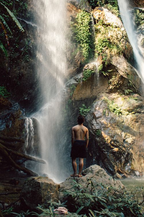 Free A man stands on a rock, admiring a waterfall surrounded by lush greenery in Chiang Mai, Thailand. Stock Photo