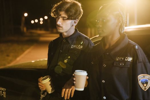 Policeman and policewoman in uniform during night patrol, holding coffee cups on a city street.
