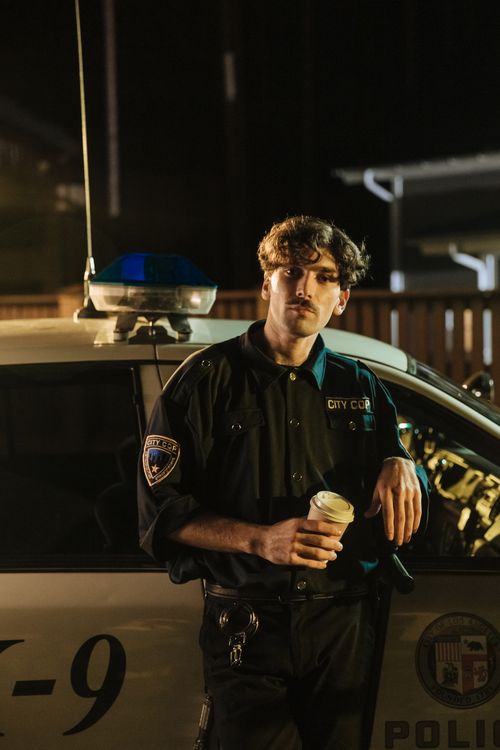 Free A police officer in uniform stands by his patrol car at night, holding a coffee cup. Stock Photo