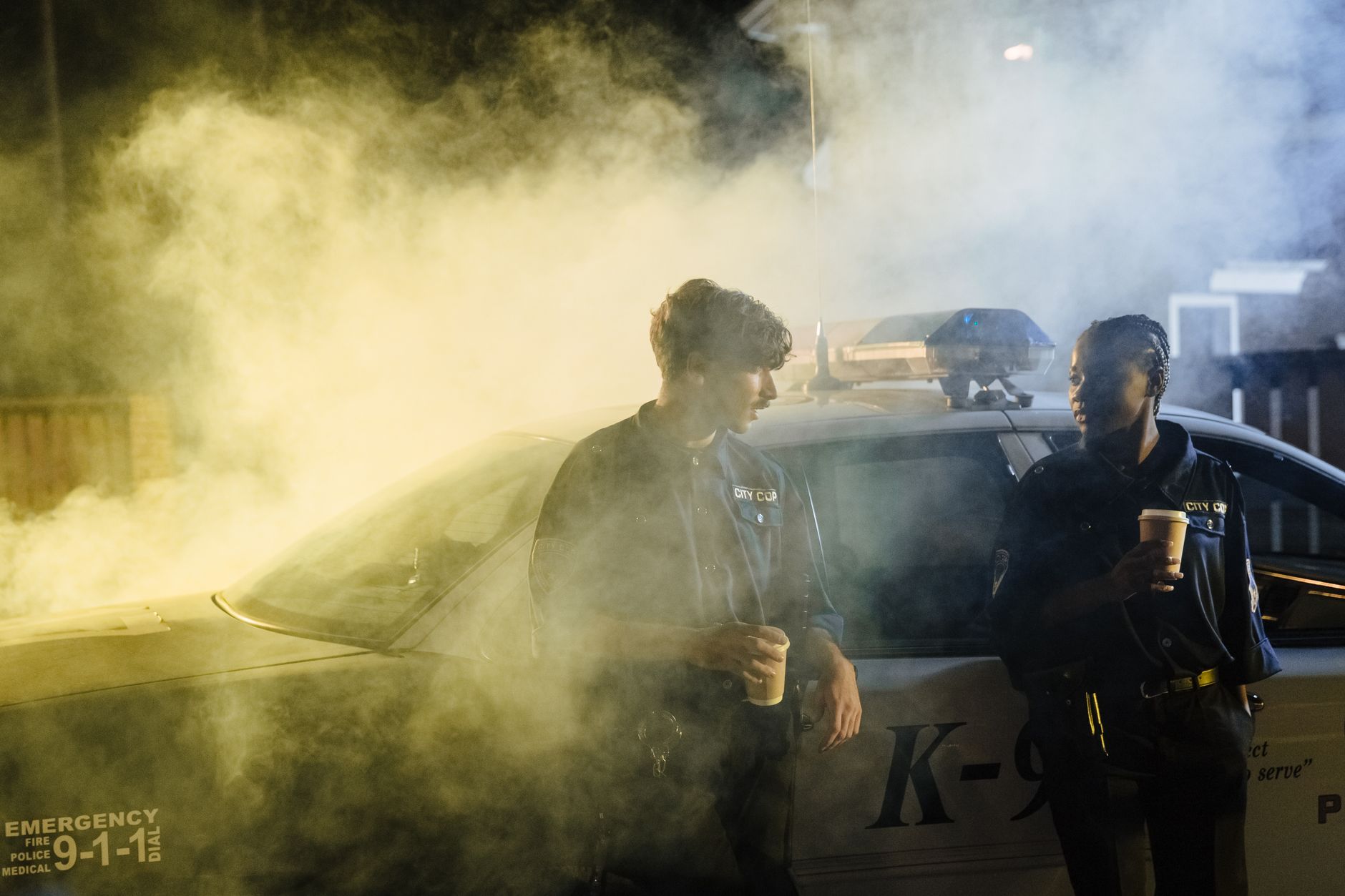 Two officers standing by a patrol car at night amidst smoke, enjoying coffee.