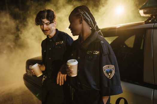 Two friendly police officers enjoying coffee beside a cop car at night.