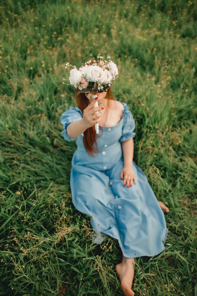 Woman In Blue Summer Dress Sitting On Meadow And Holding Bunch Of White Flowers