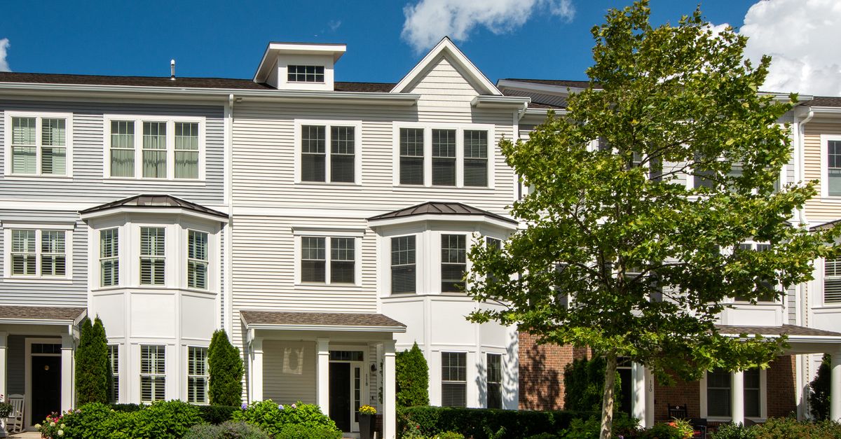 A picturesque suburban street with colonial-style homes and lush greenery under a blue sky.